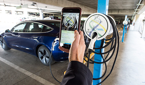 56661-float A hand holding a phone with a charging application running on it, in front of an electric vehicle and a charging station in a parking garage.